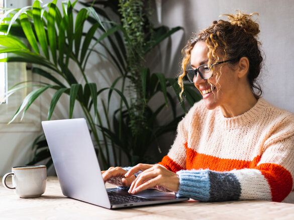 Happy woman writing on laptop on desk table in home office workplace. Adult female cheerful portrait wearing glasses and using laptop smiling. Modern alternative business job entrepreneur business, homeoffice